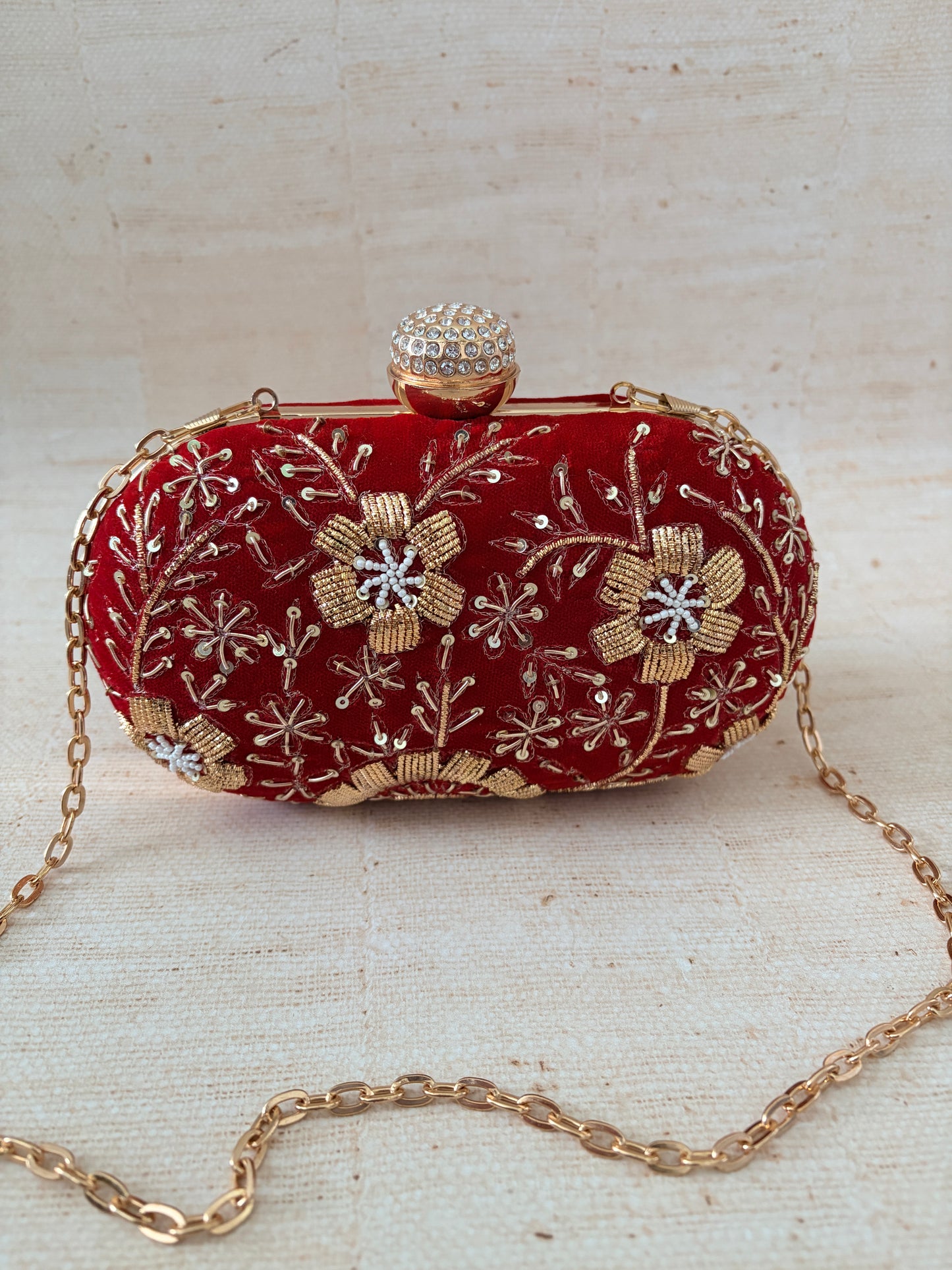 Red clutch bag with gold floral embroidery and a chain strap on a textured beige background
