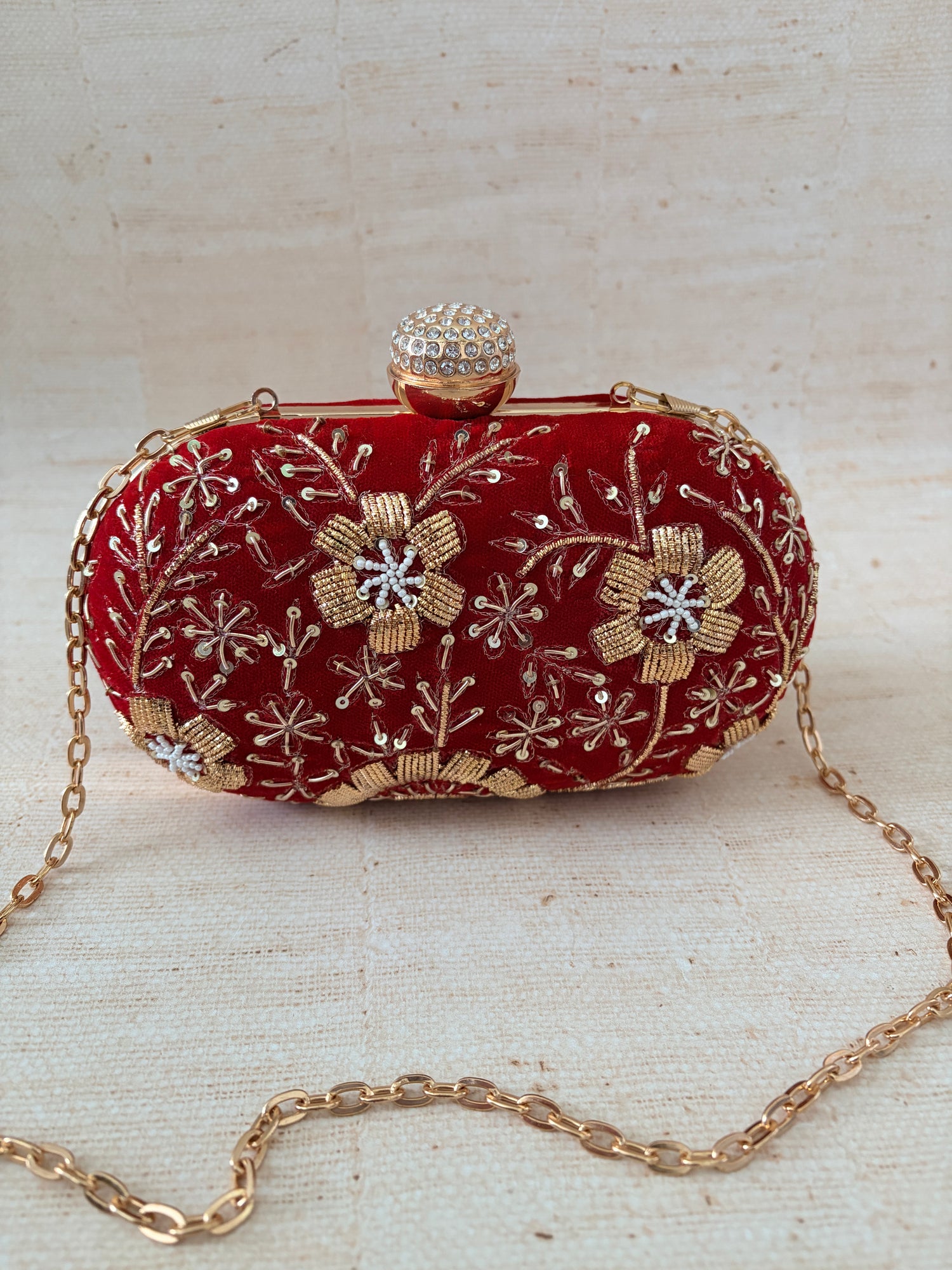Red clutch bag with gold floral embroidery and a chain strap on a textured beige background