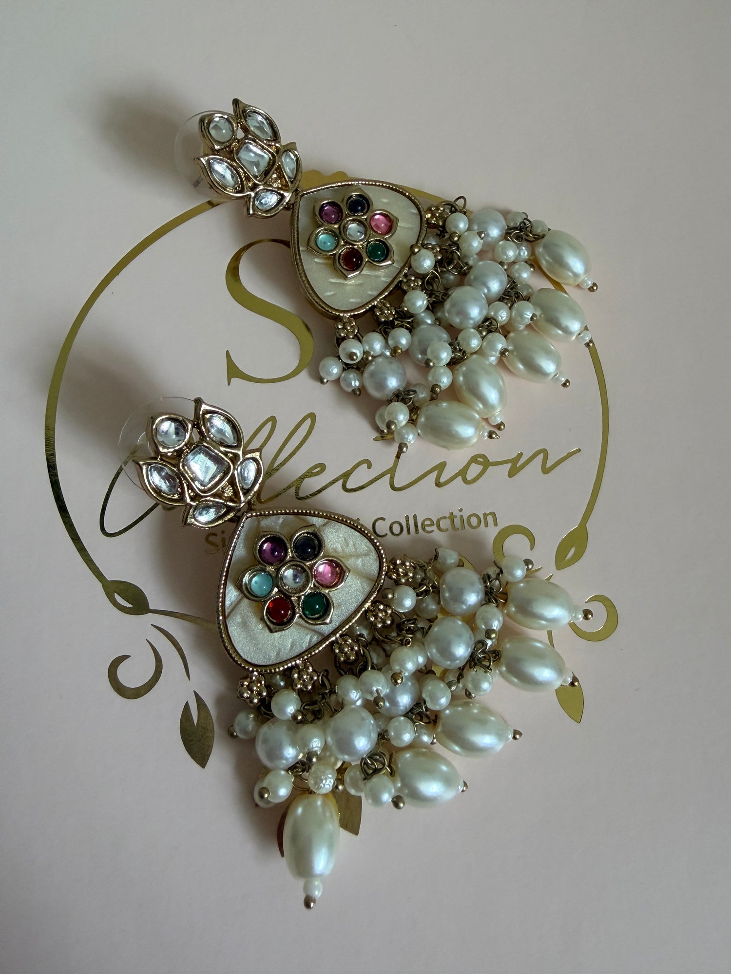 Decorative brooch with pearls and gemstones on a pink background with 'SR Collection' branding.