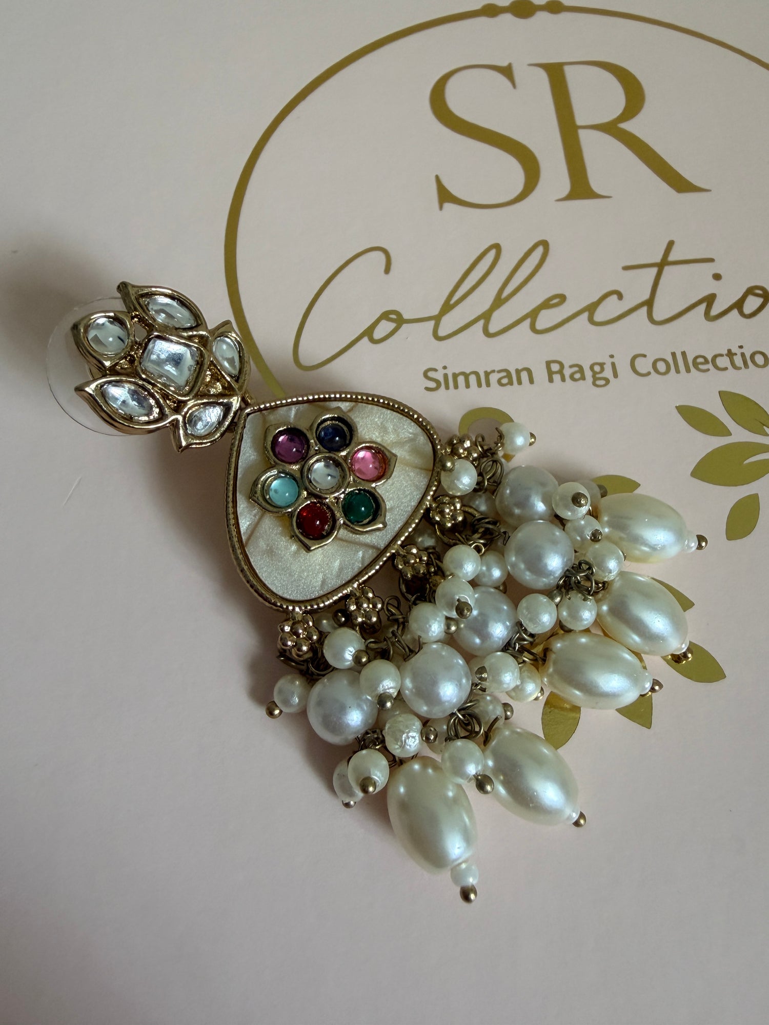 Decorative brooch with pearls and gemstones on a pink background with 'SR Collection' branding.