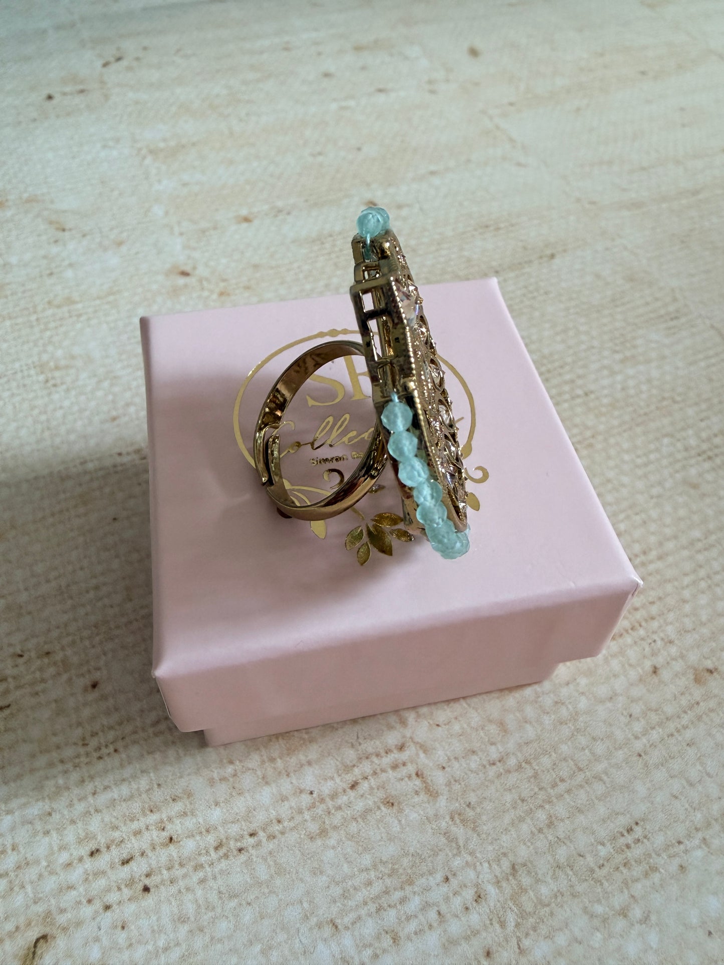 Decorative ring with gemstones on a pink box against a textured beige background
