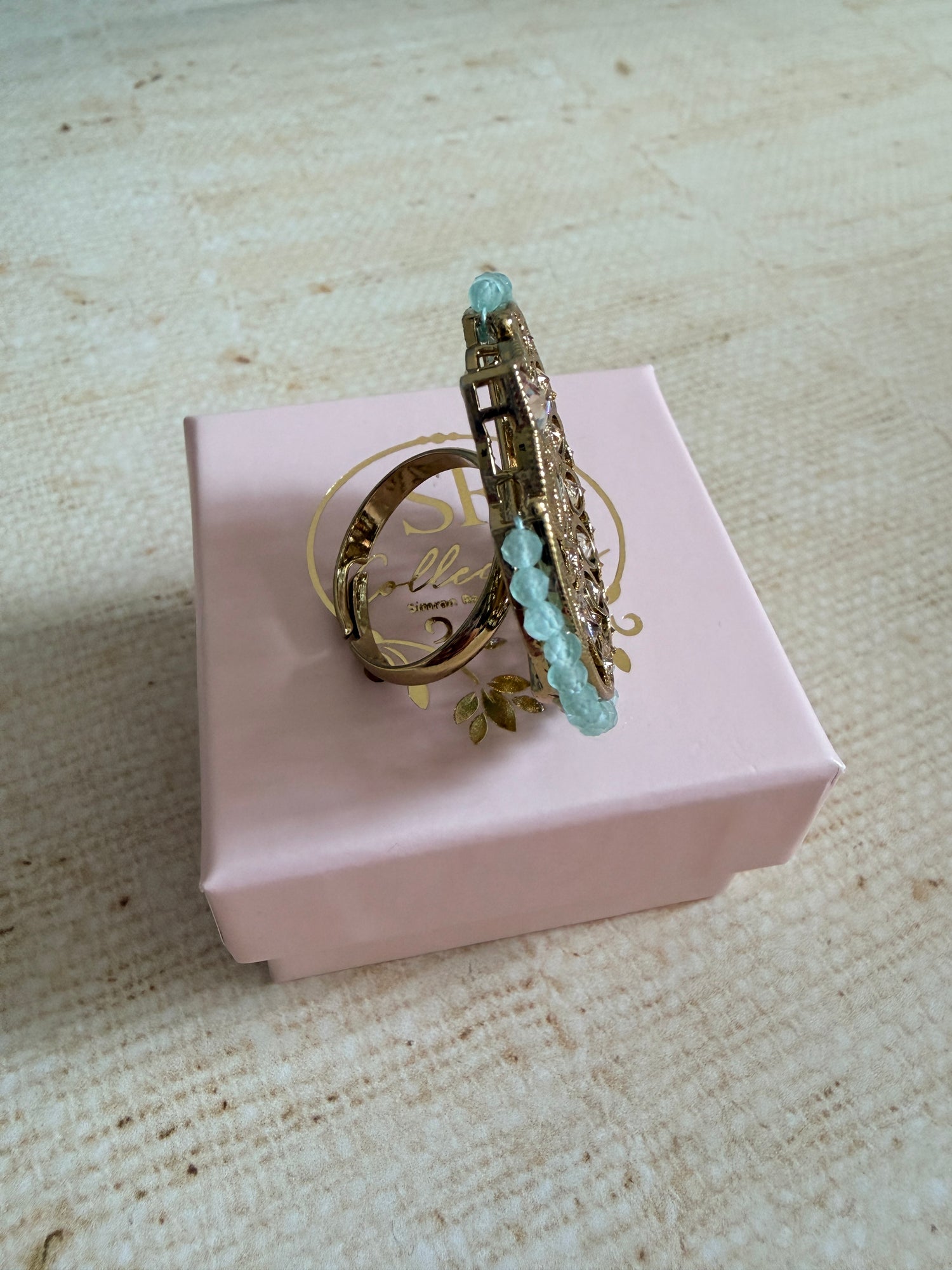 Decorative ring with gemstones on a pink box against a textured beige background