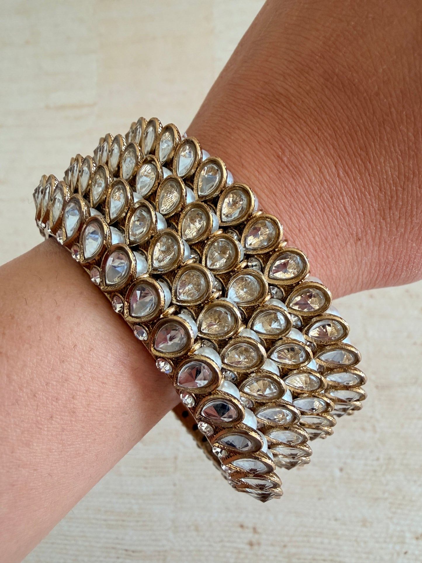 Close-up of a person wearing a stack of ornate gold and gemstone bracelets on a neutral background.
