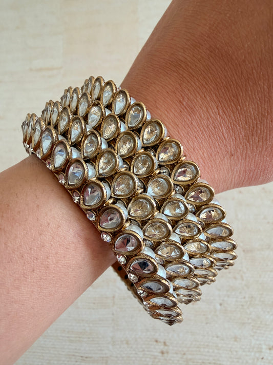 Close-up of a person wearing a stack of ornate gold and gemstone bracelets on a neutral background.